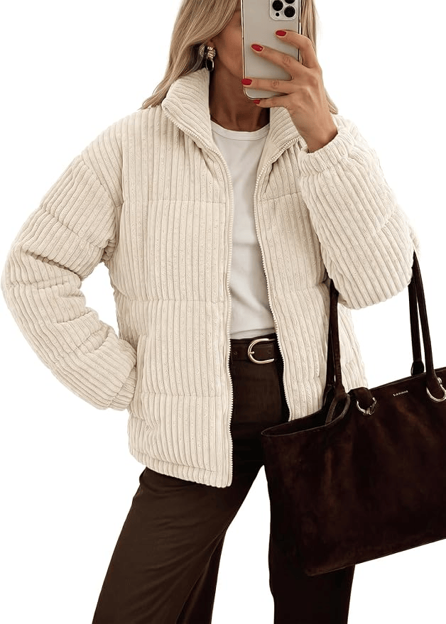 A woman wearing a cream-colored puffer jacket, white shirt, and brown pants, holding a dark handbag with one hand and taking a selfie with the other.