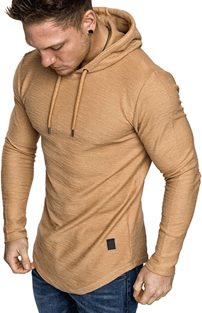 A man wearing a tan long-sleeve hoodie, looking down thoughtfully while adjusting the hoodie. He has short hair and is dressed in dark jeans.