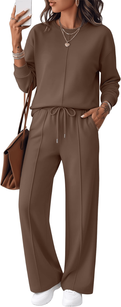A woman wearing a matching brown loungewear set consisting of a long-sleeve top and loose pants, paired with white sneakers and accessorized with layered necklaces and bracelets. She is holding a smartphone and has a brown handbag slung over her shoulder.