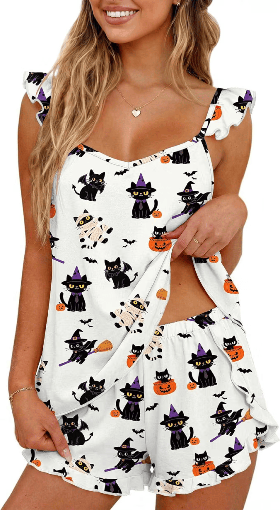 A woman wearing a cute, Halloween-themed pajama set featuring black cats, pumpkins, and bats, smiling cheerfully.