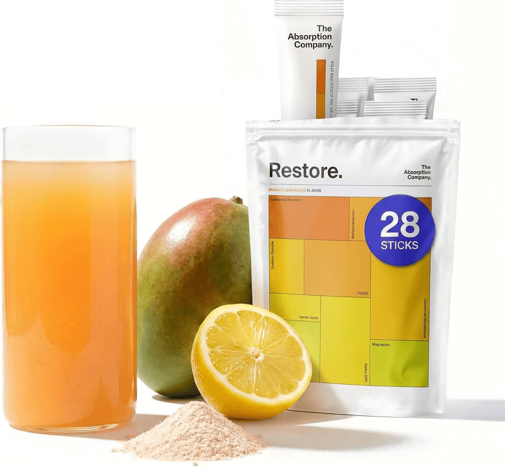 A glass of orange beverage next to a mango, a halved lemon, and a pouch of Restore powdered drink mix featuring its label and packaging.